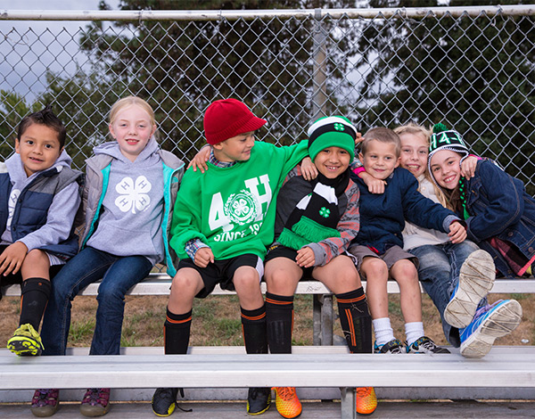 seven 4-H children sitting on a bench outdoors with chain link fence in background