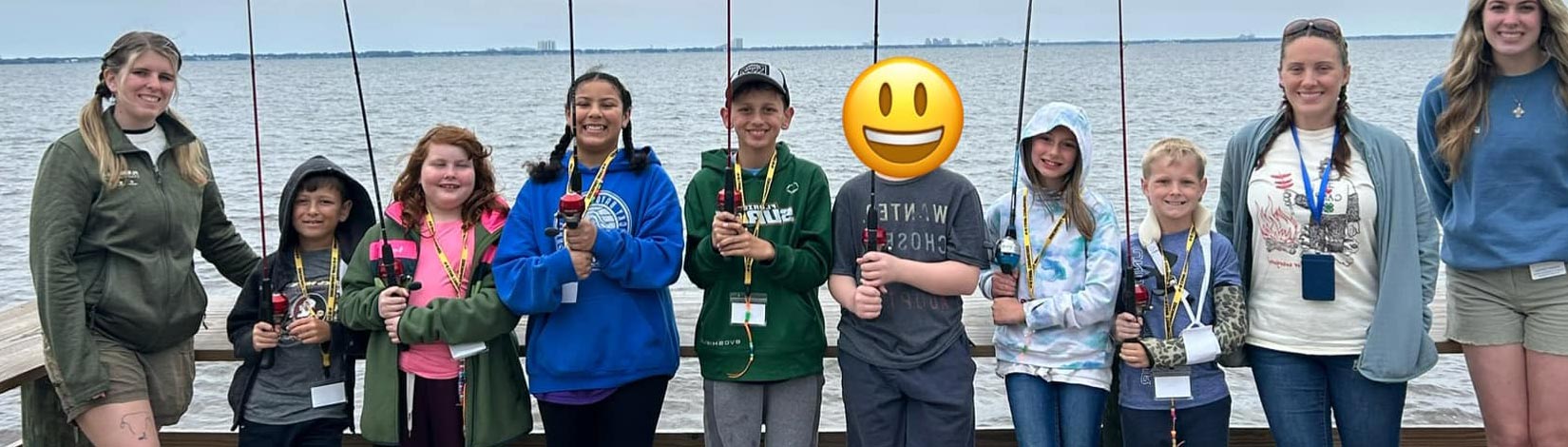 A group of kids with fishing poles, accompanied by camp counselors, stand on a dock at Camp Timpoochee, ready to fish.