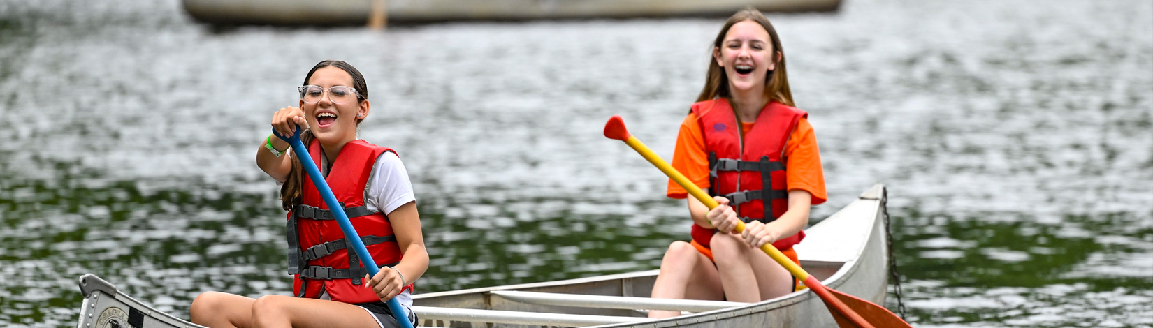 Two smiling young girls wearing life vests paddle a canoe across a calm lake on a sunny day.