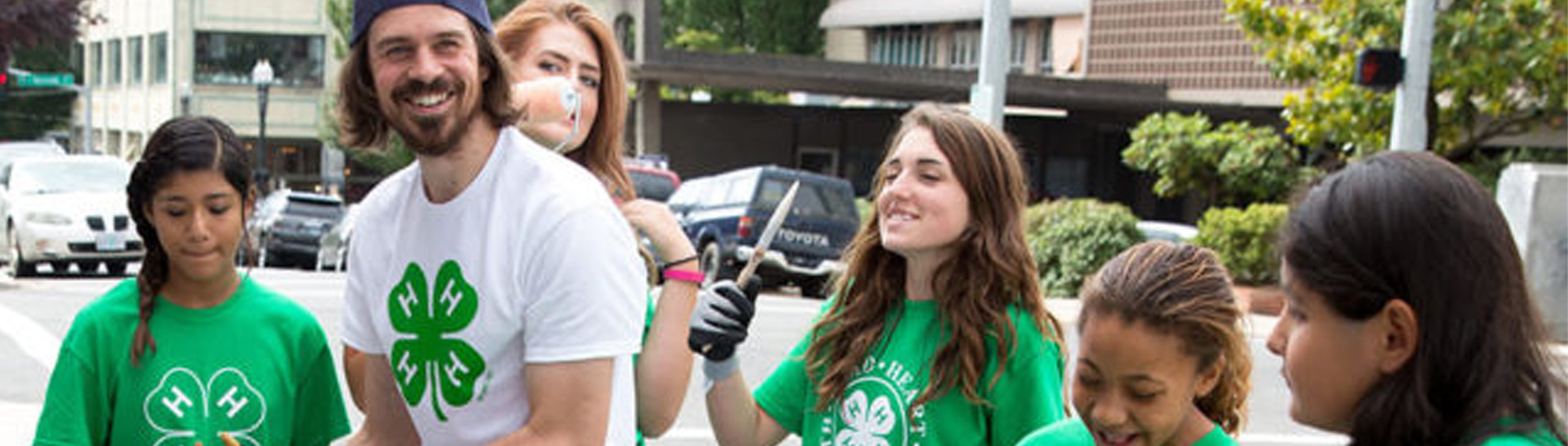 A volunteer artist with a warm smile stands outside in an urban setting, surrounded by a group of kids with paint supplies.