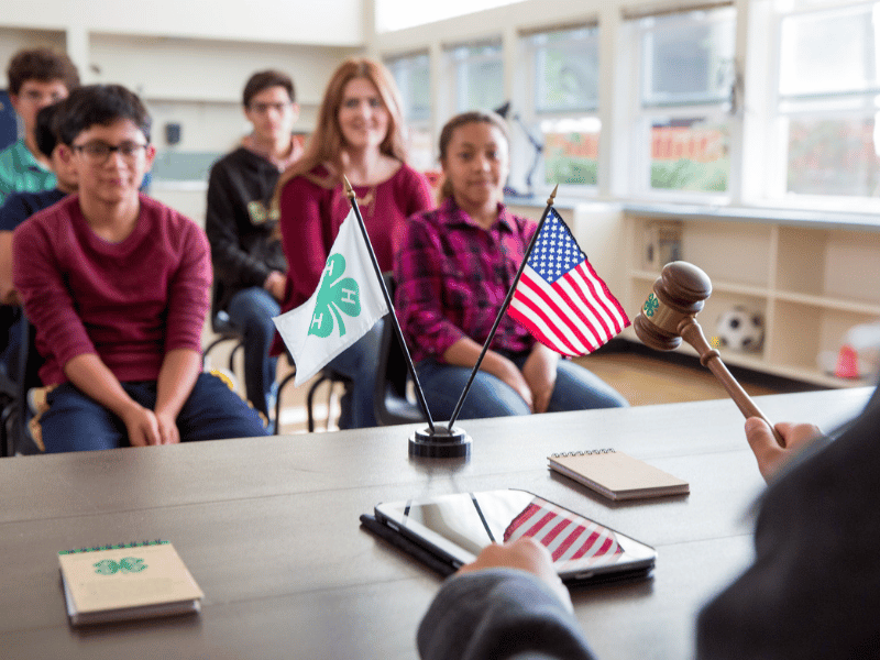 Six children sitting in a classroom, a person off camera holds a gavel.