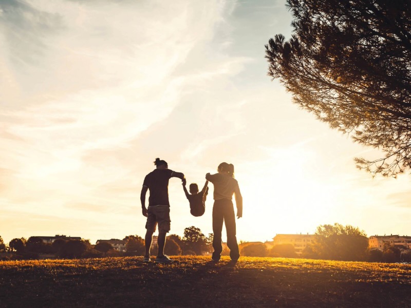 Silhouette of two adults swinging a child between them at sunset in a park.