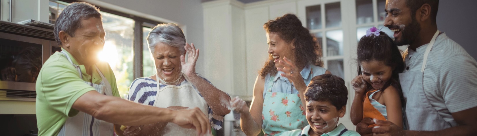 A multigenerational family in aprons joyfully baking together in a bright home kitchen, tossing flour and stirring a bowl near an oven and white cabinets; a man holds a girl with a purple flower in her hair while a boy looks on.