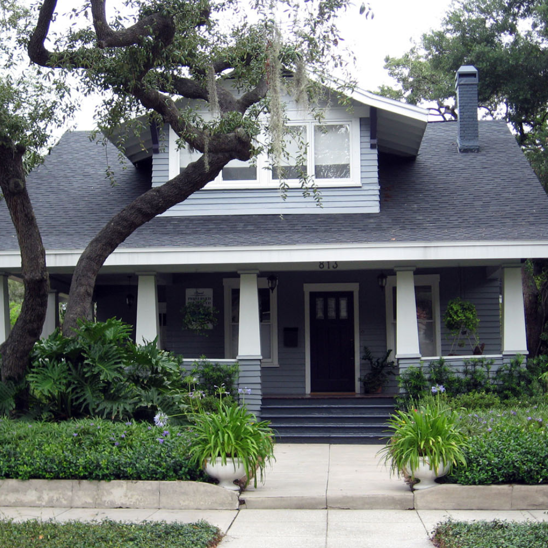 House with gabled roof and front porch surrounded by trees, bushes, and a concrete walkway.