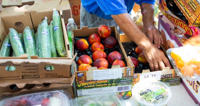 photo of a vegetable stand with a person wearing a blue shirt placing vegetables on table. There are green cucumbers, peaches, and boxes of fruit and veg showing on table.