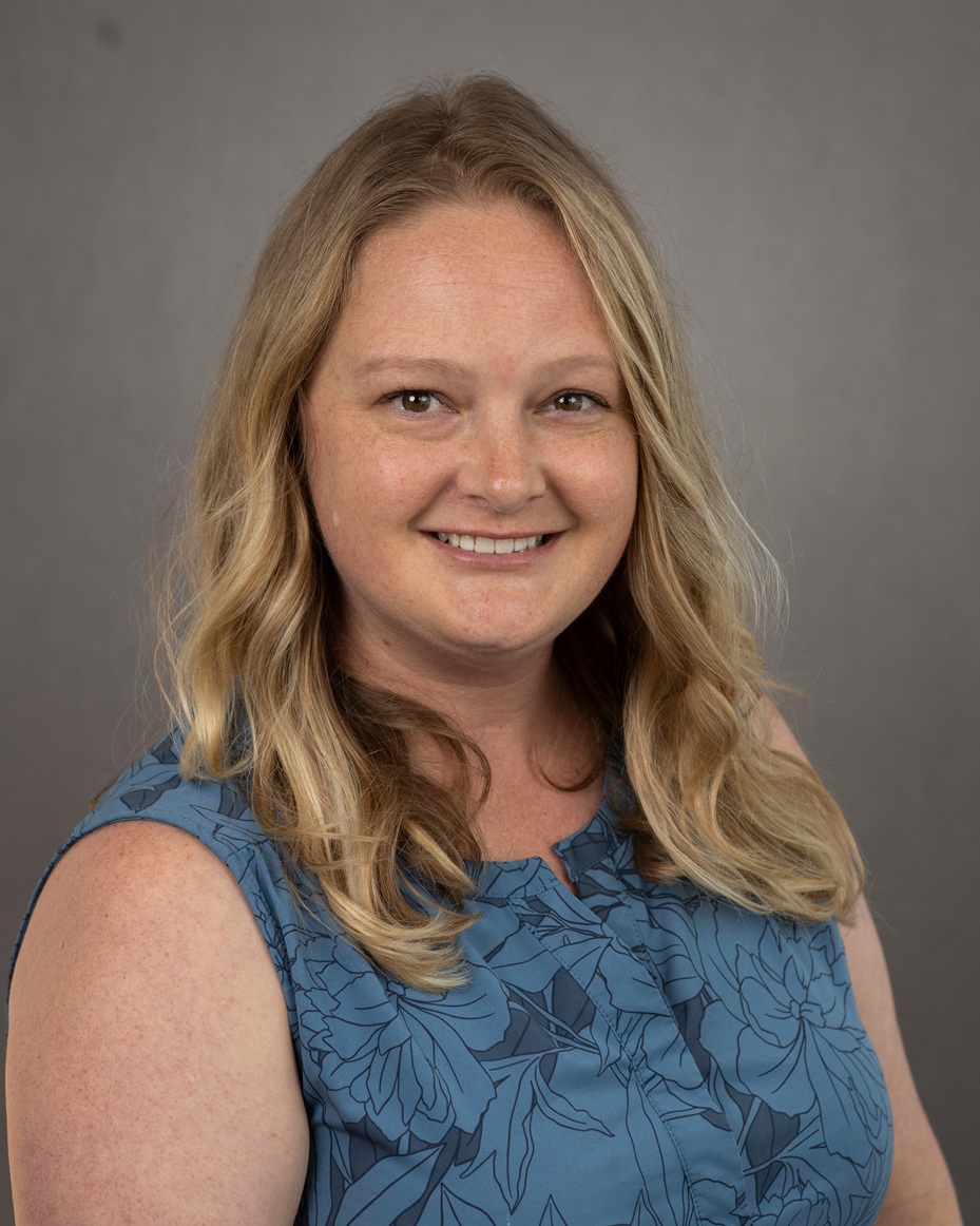 A professional headshot of woman with long blonde hair, wearing a blue sleeveless dress in front of a gray background.