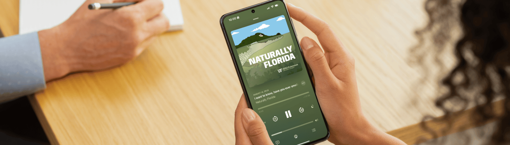 Two people sit at a wood table which shows the cellphone of one is playing the Naturally Florida podcast.