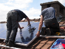 two workers install solar power panels near the peak of a red-tiled rooftop