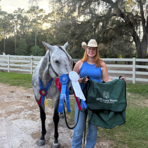 Person in a cowboy hat holding a green '2023 Area North 4-H Horse Show' bag next to a gray horse adorned with award ribbons, standing near a white fence and trees
