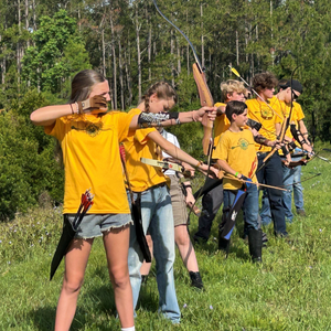 Group of youth in yellow T-shirts practicing archery on a grassy field with tall trees in the background