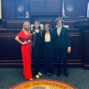 Four individuals in professional attire standing in a formal government chamber with the Florida state seal, U.S. and Florida flags, and a large wooden podium in the background